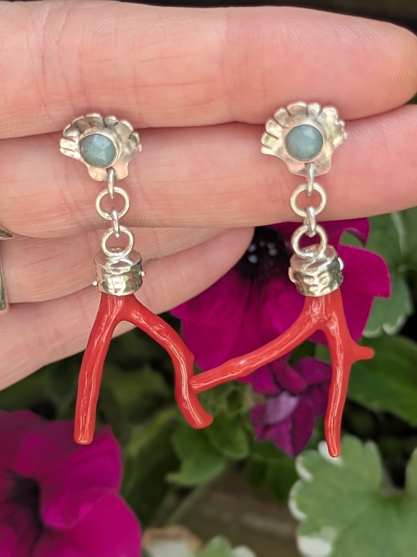 A close-up of handmade earrings resting on a hand with silver rings. The earrings have silver scallop shell studs with turquoise gemstones and dangling red coral branch drops. Background includes deep green leaves and purple-pink flowers, enhancing the natural, boho style.
