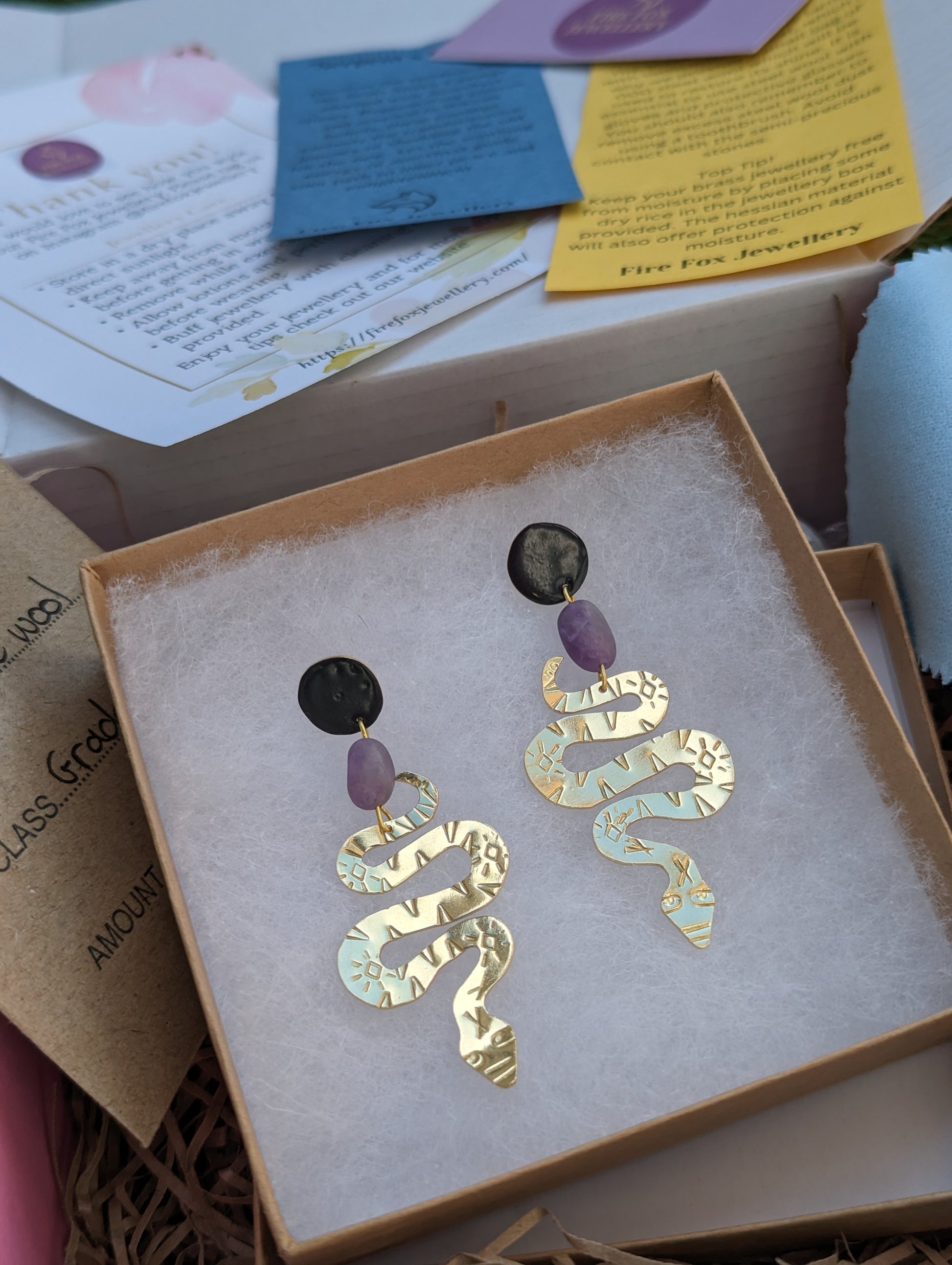 Brass textured snake earrings with amethyst beads hanging from black enamel brass studs. The earrings are placed in an eco friendly jewellery box with information leaflets in the background.