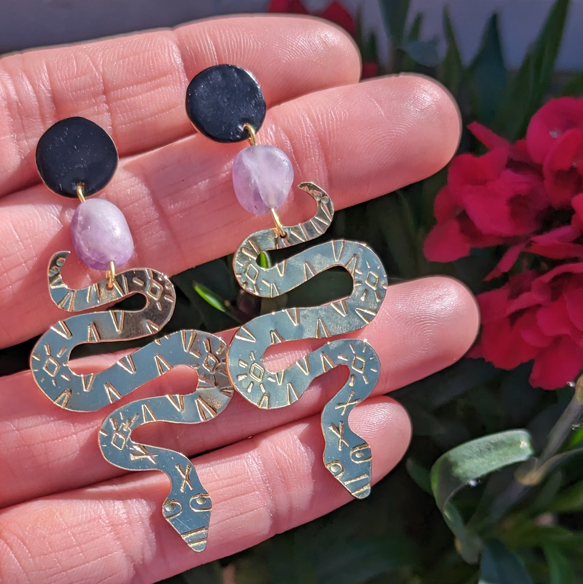 Brass textured snake earrings with amethyst beads hanging from black enamel brass studs. The earrings are placed on a hand to show the contrast with skin with a floral background.