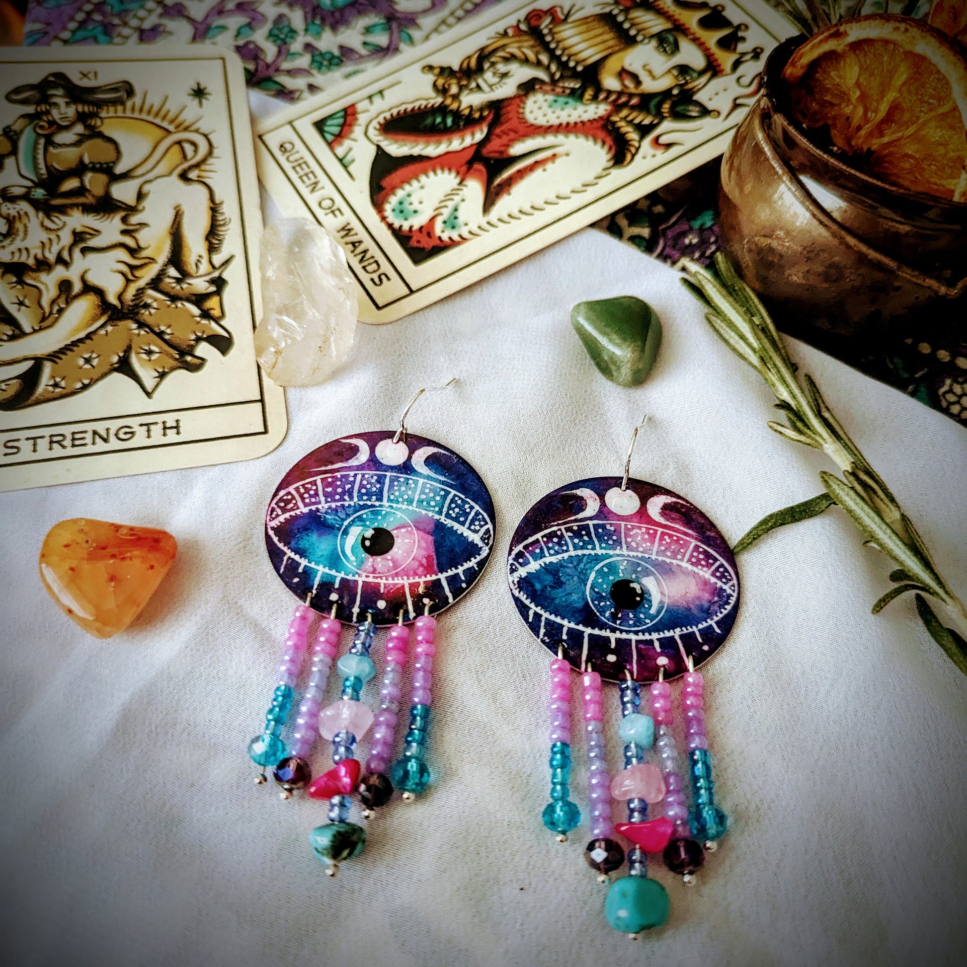 Earrings. Large circular aluminium disc enamelled with a mystical colour combination with a large eye in the centre with a crescent moons above the eyelid. Five silver plated pins hang from the eye, embellished with acrylic beads, glass crystals and healing crystals. including  rose quartz, turquoise, pink howlite and aquamarine. The earrings are displayed with wicca items including cauldron and tarot cards 