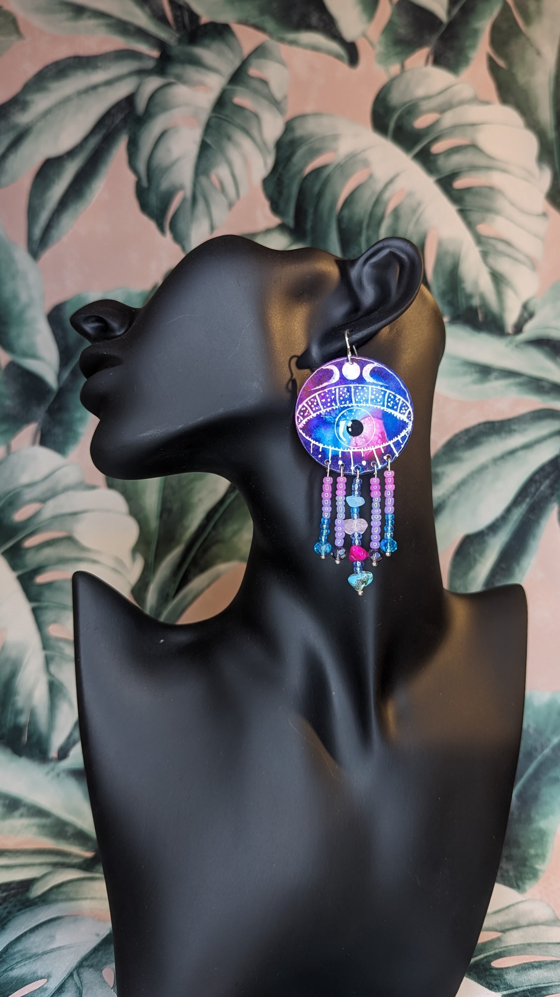 A single earring. large circular aluminium disc enamelled with a mystical colour combination with a large eye in the centre with a crescent moons above the eyelid. Five silver plated pins hang from the eye, embellished with acrylic beads, glass crystals and healing crystals. including rose quartz, turquoise, pink howlite and aquamarine. The earring is displayed on a black mannequin with a floral background 