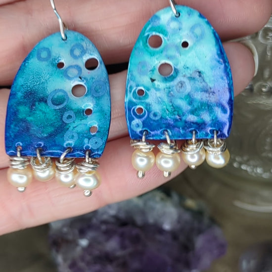 Video of blue enamel earring adorned with apricot freshwater pearls, placed on a hand, the earrings are moving to show all the blue colours 