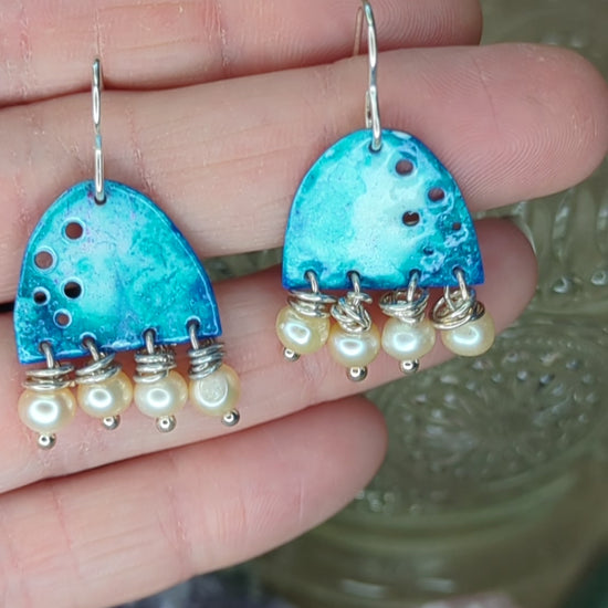 Video of blue enamel earring adorned with apricot freshwater pearls, placed on a hand, the earrings are moving to show all the blue colours 