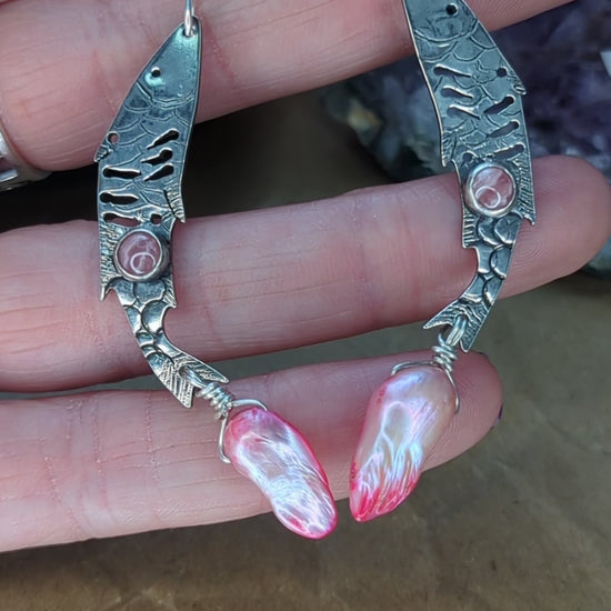 Sterling silver fish earrings, with fish details engraved on the silver, adorned with pink Rhodochrosite Cabochons and Pink keshi Freshwater Pearls. The earrings are placed on a hand, the earrings are moving around to show the detail on the fish earrings. 