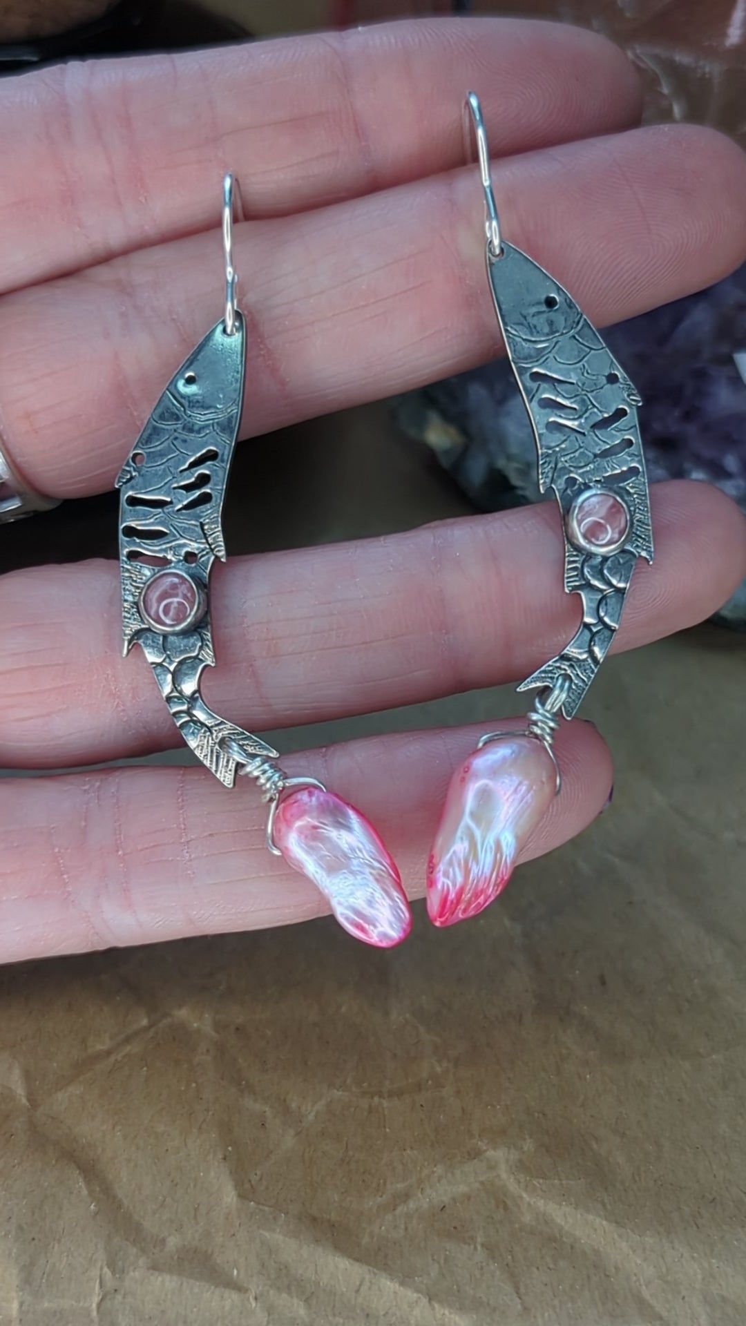 Sterling silver fish earrings, with fish details engraved on the silver, adorned with pink Rhodochrosite Cabochons and Pink keshi Freshwater Pearls. The earrings are placed on a hand, the earrings are moving around to show the detail on the fish earrings. 