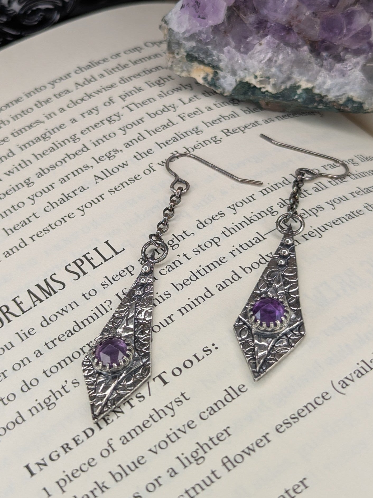 Maiden Earrings placed on an open spell book, with a raw amethyst stone in the background, creating a mystical and enchanting atmosphere.

