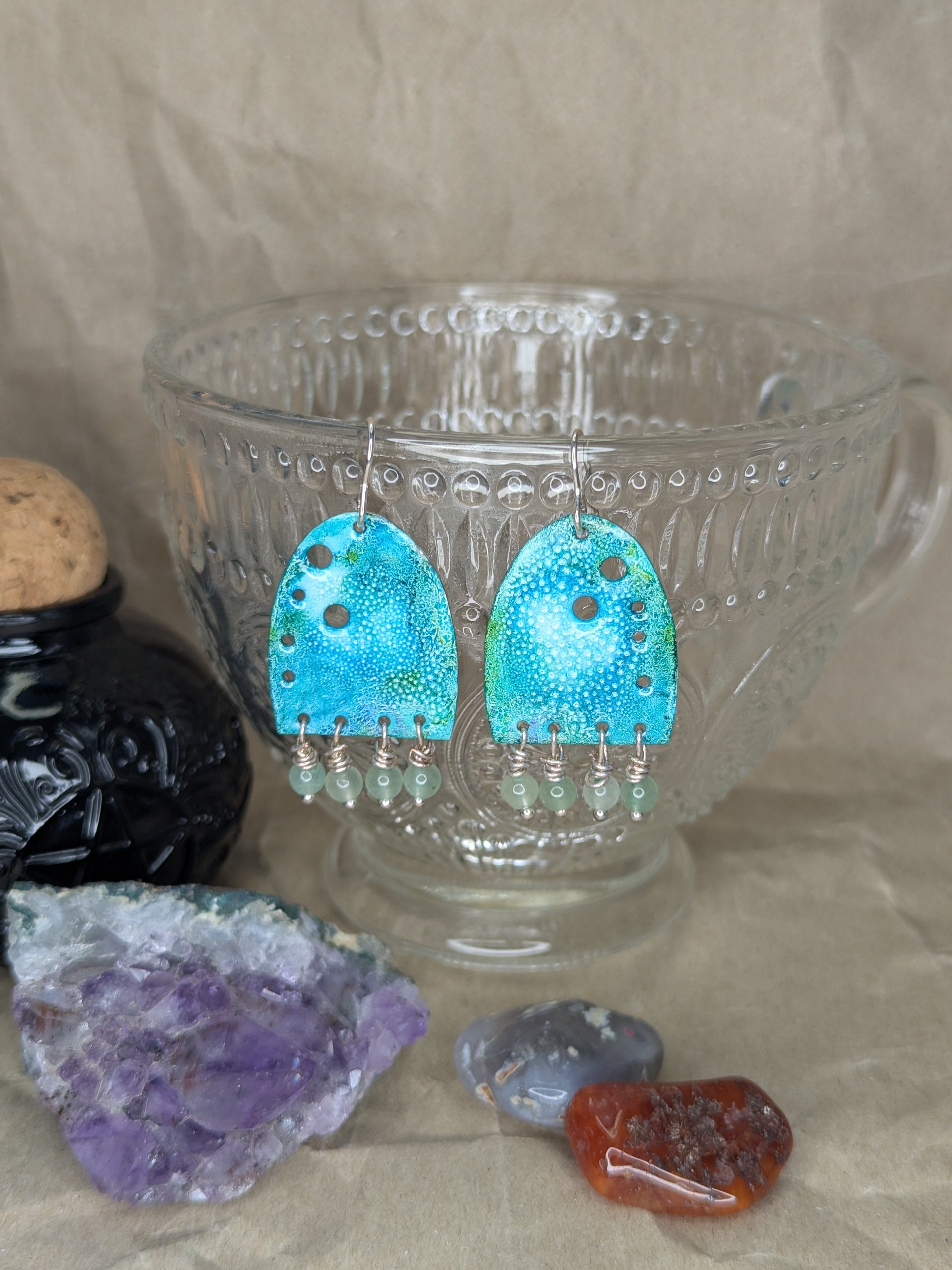 Handcrafted turquoise enamel earrings with colours of blue and turquoise adorned with green aventurine beads. The earrings are hanging from a crystal teacup