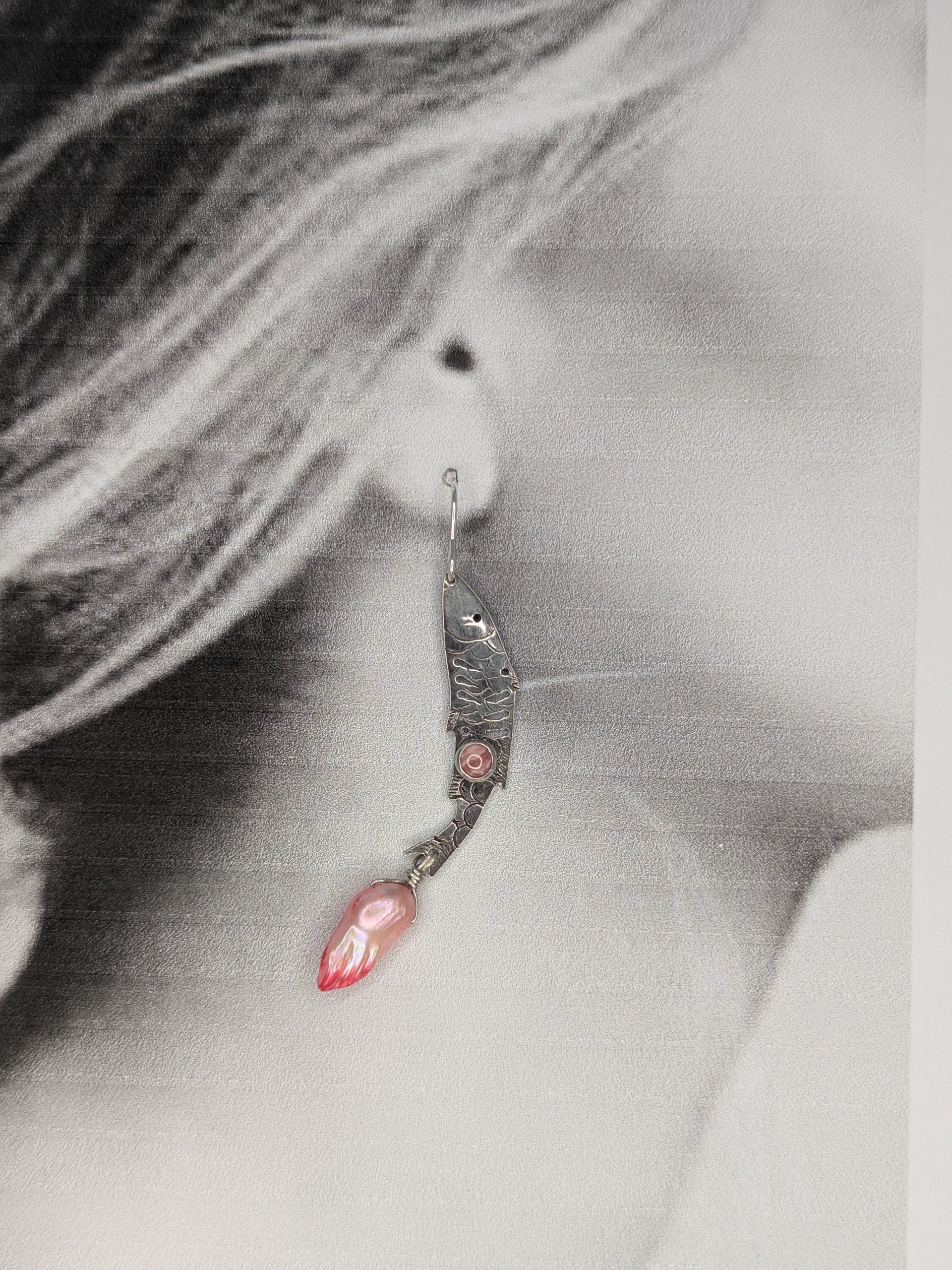 Sterling silver fish earrings, with fish details engraved on the silver, adorned with pink Rhodochrosite Cabochons and Pink keshi Freshwater Pearls. The earrings are placed on a black and white image of a model to show the earrings in the ear lobe. 