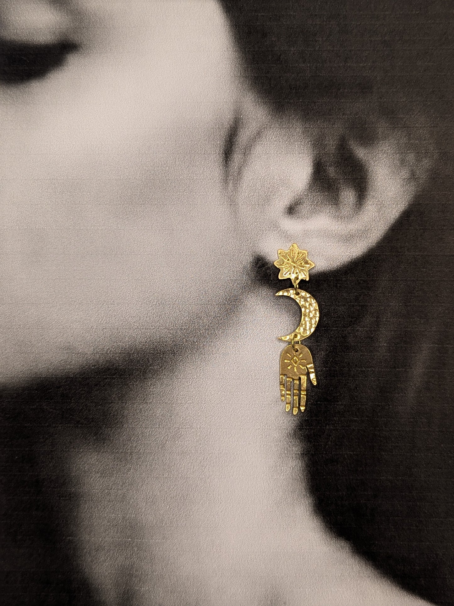Gold brass earrings are hanging from a models ears, to enhance the beautiful detail of each component and show how beautiful they look while wearing them. Hand-cut from raw gold brass, featuring a star stud, a crescent moon, and a protective hand delicately dangling below.