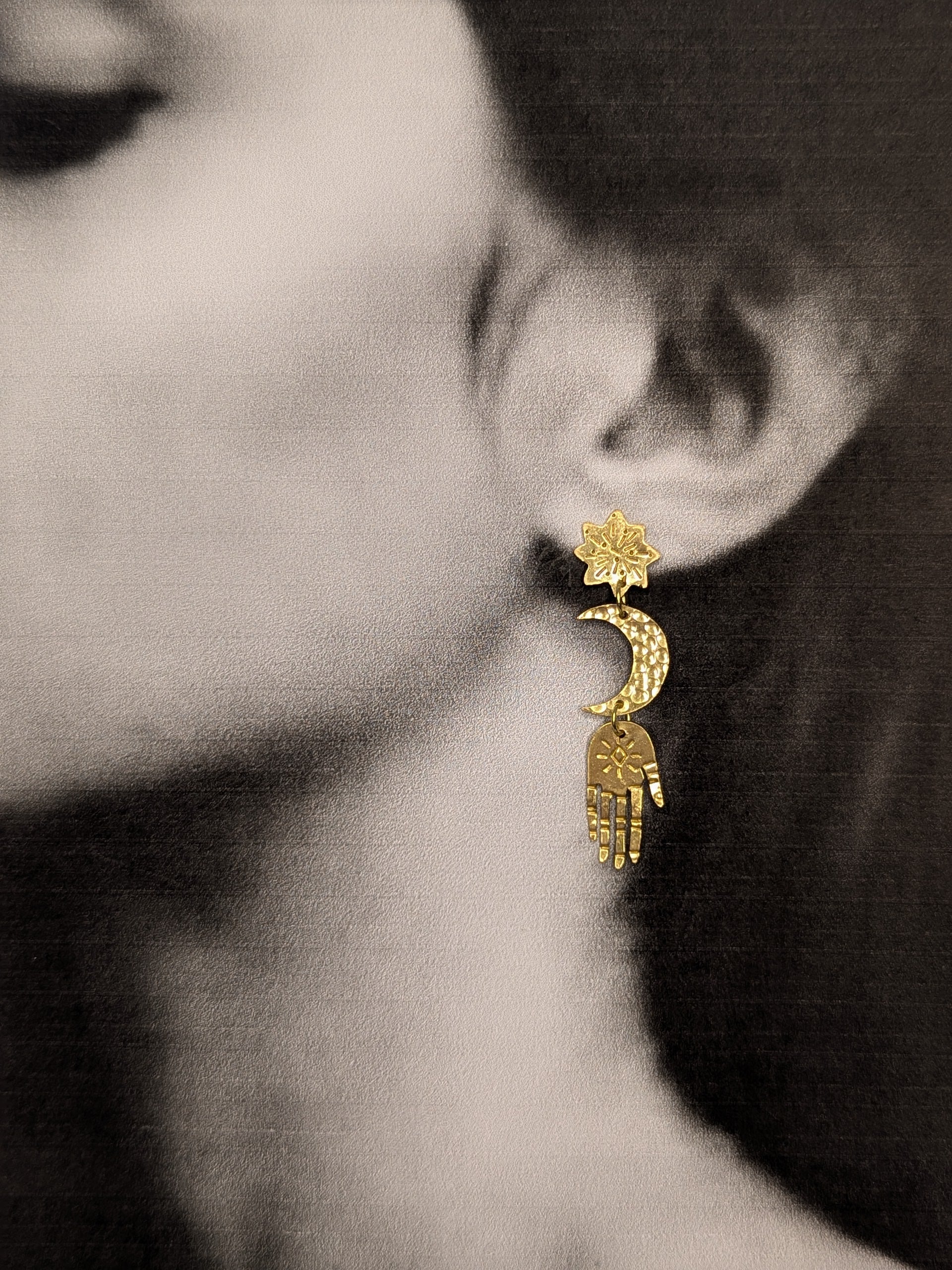 Gold brass earrings are hanging from a models ears, to enhance the beautiful detail of each component and show how beautiful they look while wearing them. Hand-cut from raw gold brass, featuring a star stud, a crescent moon, and a protective hand delicately dangling below.