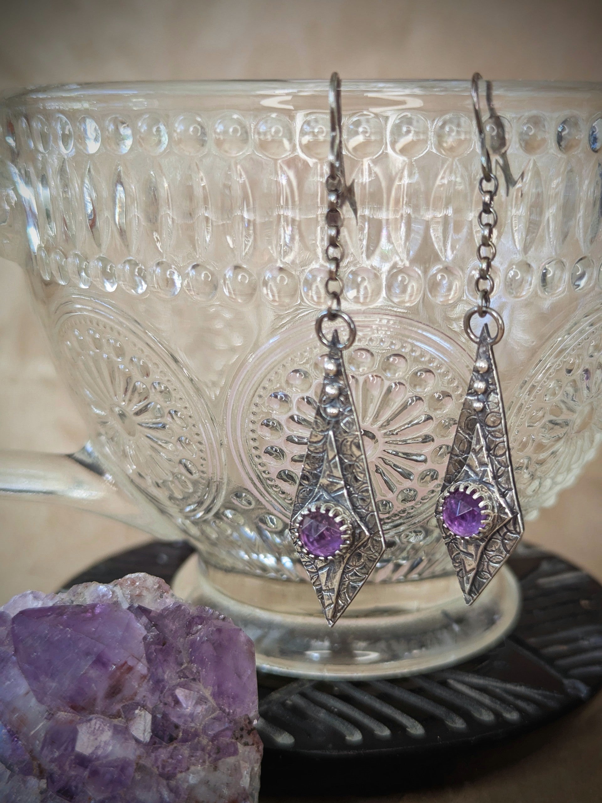 Maiden Earrings hanging gracefully from the rim of a delicate crystal teacup, accompanied by a sparkling amethyst stone beside the cup, blending elegance with spiritual charm.
