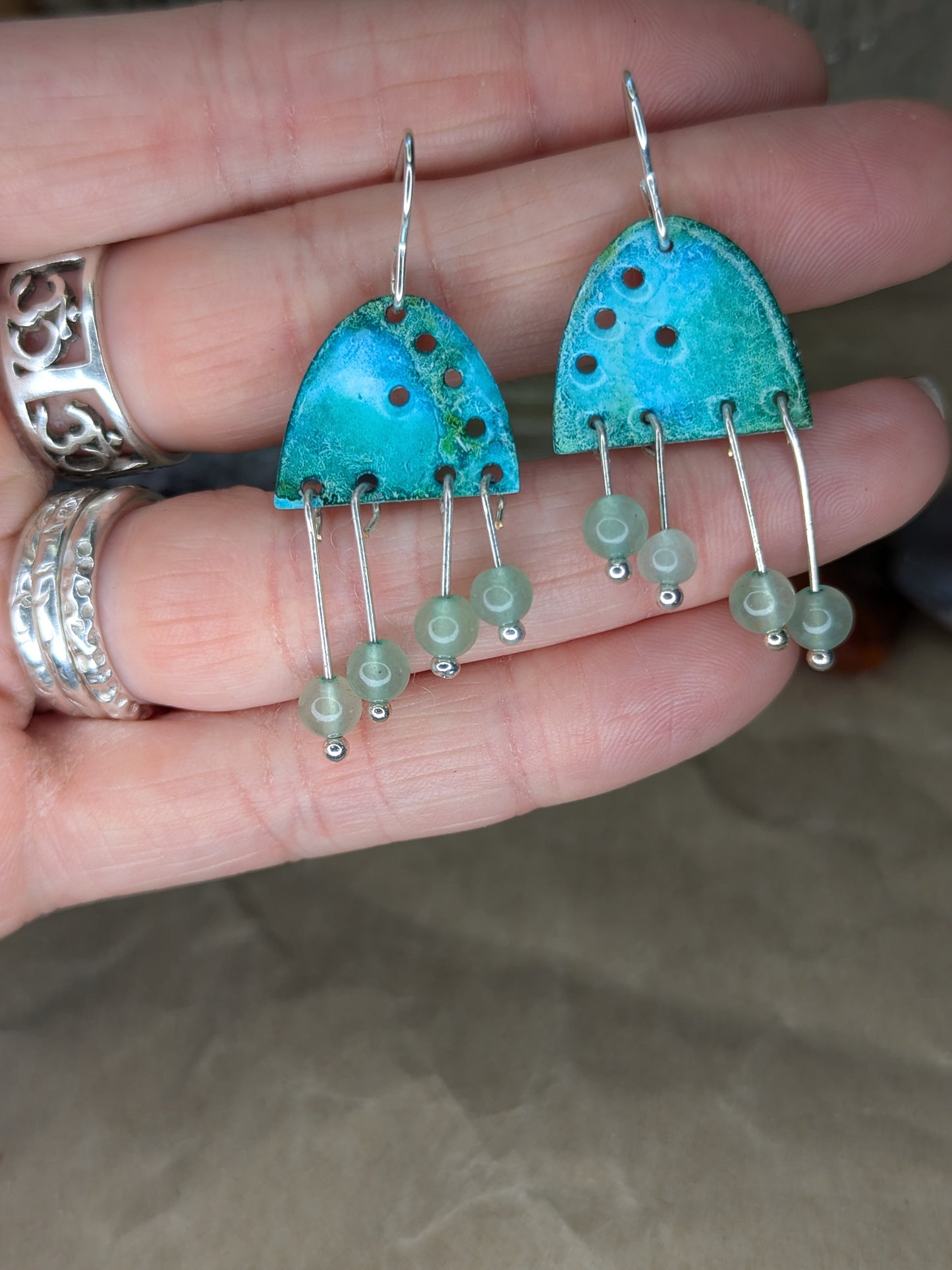 Turquoise enamel earrings with colours of blue and turquoise adorned with green aventurine beads, Earrings are placed on hands to show the contrast with the earrings against skin.