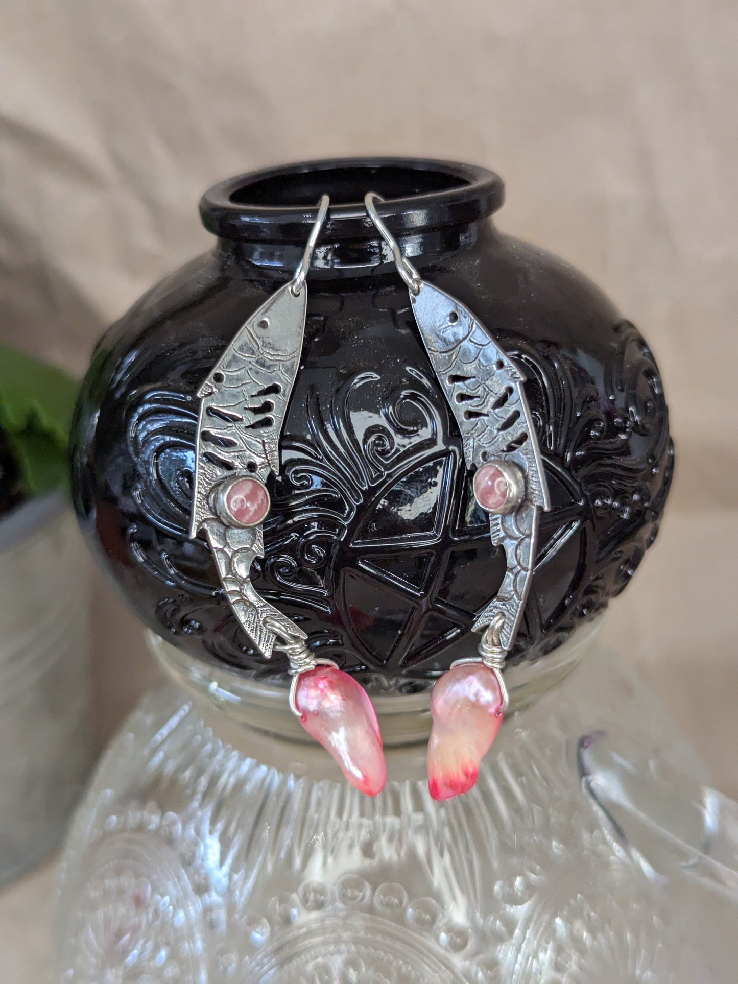 Sterling silver fish earrings, with fish details engraved on the silver, adorned with pink Rhodochrosite Cabochons and Pink keshi Freshwater Pearls. The earrings are placed on a black spell jar hanging from the neck of the jar. The spell jar is placed on a crystal glass cup with brown packing paper in the background. 