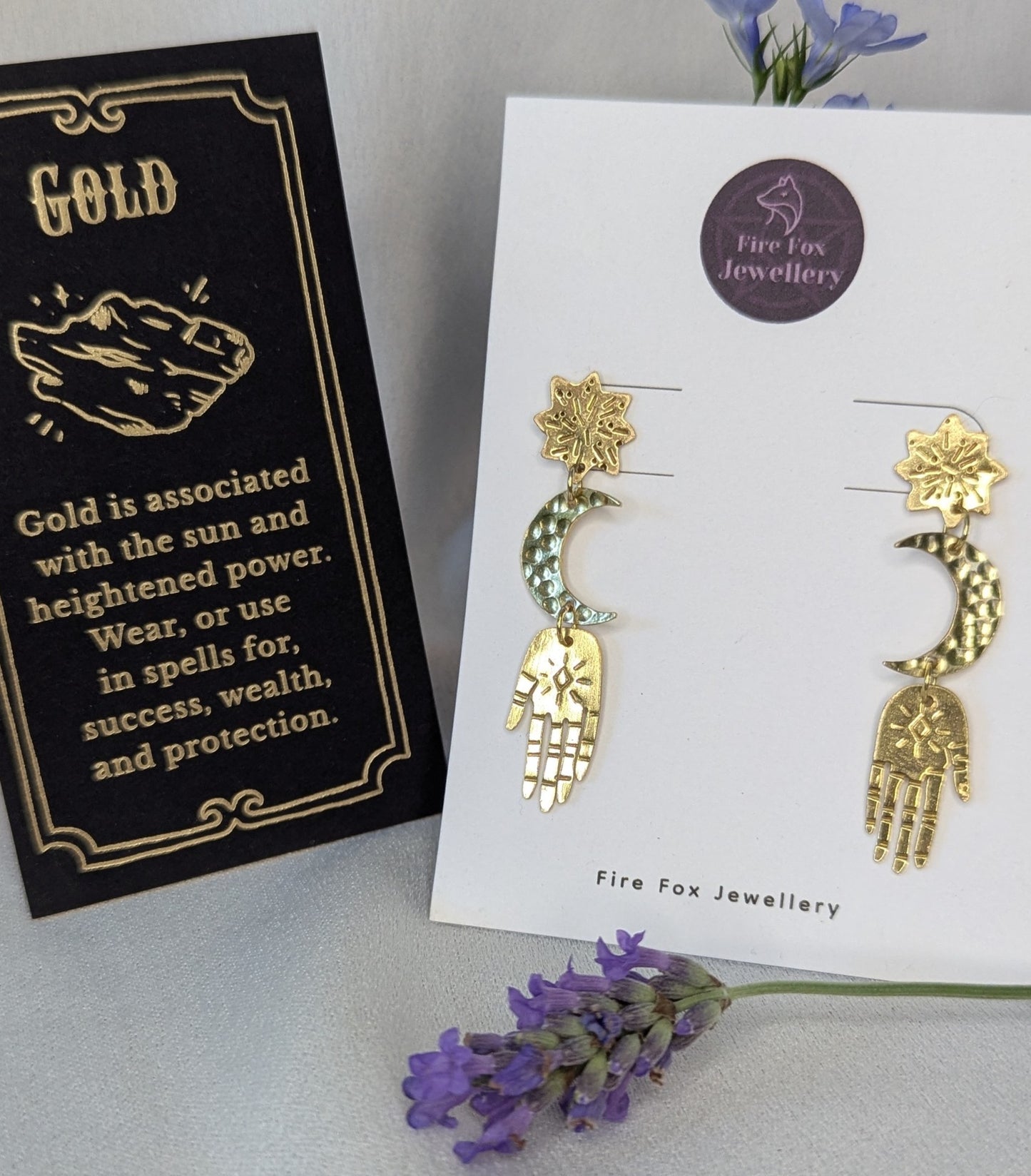 Gold brass earrings placed on a white Fire Fox Jewellery display card to enhance the beautiful detail of each component. Hand-cut from raw gold brass, featuring a star stud, a crescent moon, and a protective hand delicately dangling below. In the background there is purple flowers next to a card that details the symbolism of wearing gold which is success, wealth and protection.