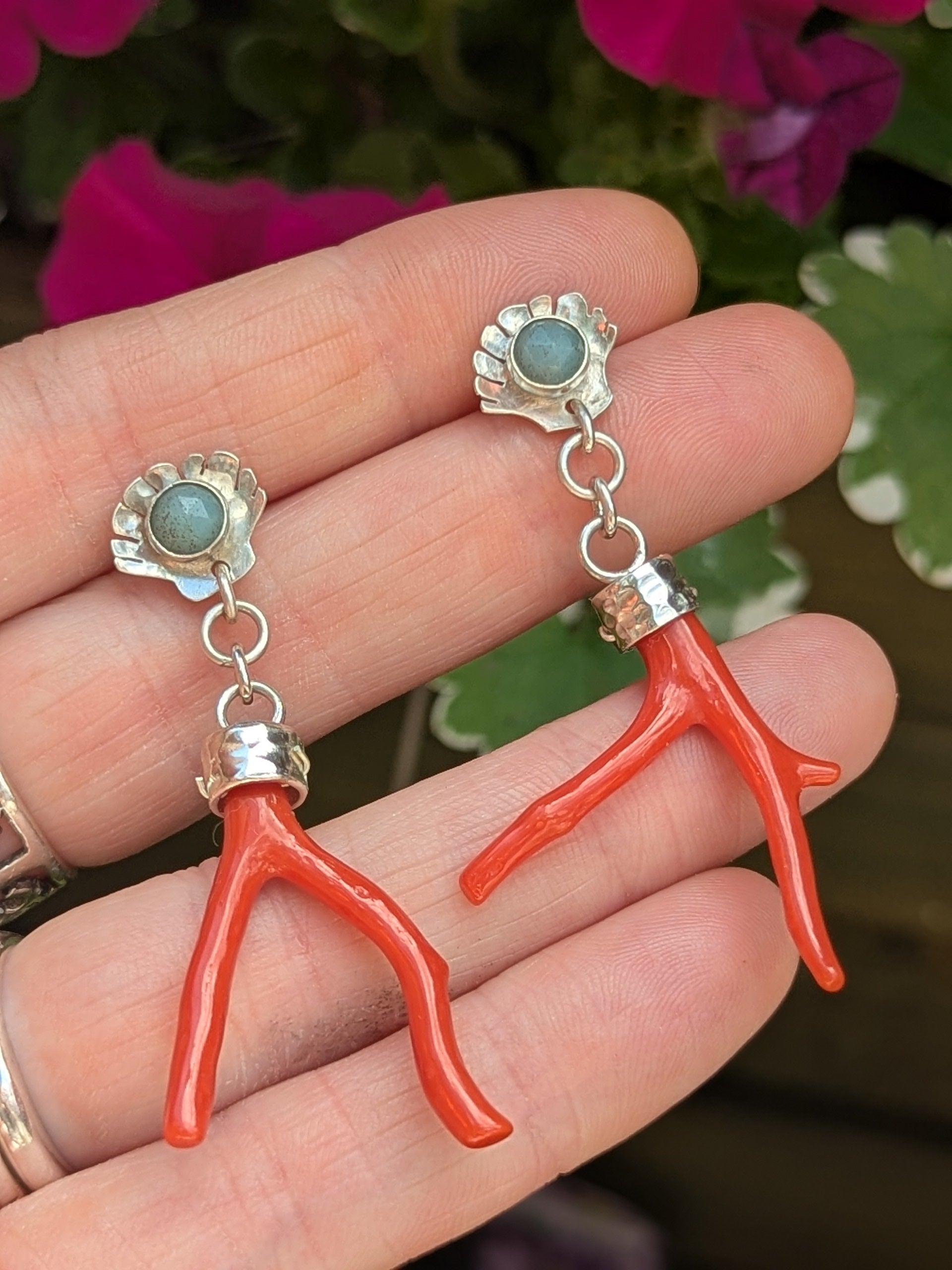 A close-up of handmade earrings resting on a hand with silver rings. The earrings have silver scallop shell studs with turquoise gemstones and dangling red coral branch drops. Background includes deep green leaves and purple-pink flowers, enhancing the natural, boho style.