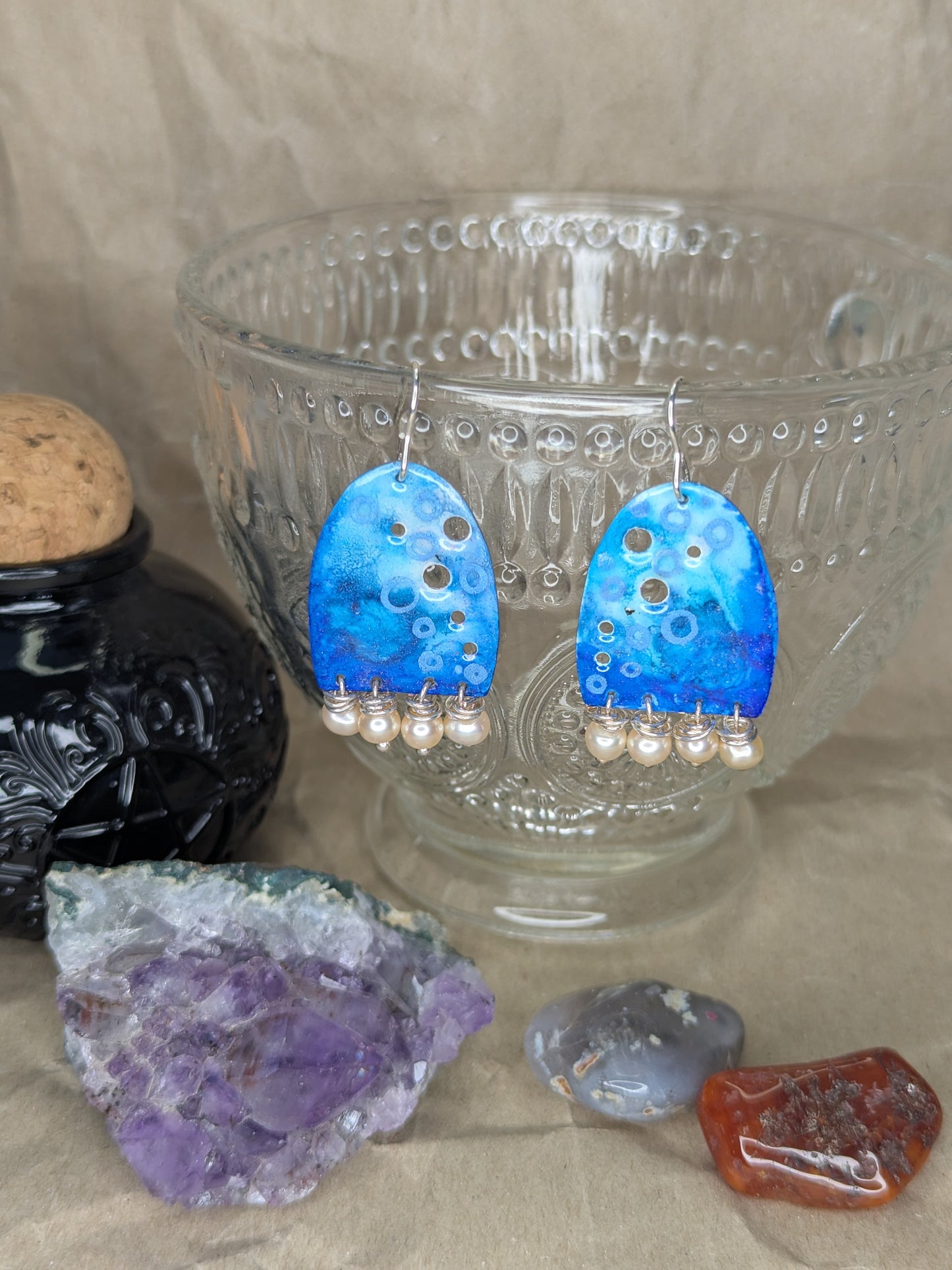 Handcrafted blue enamel earrings with colours of blue adorned with apricot freshwater pearls. The earrings are hanging from a crystal teacup.