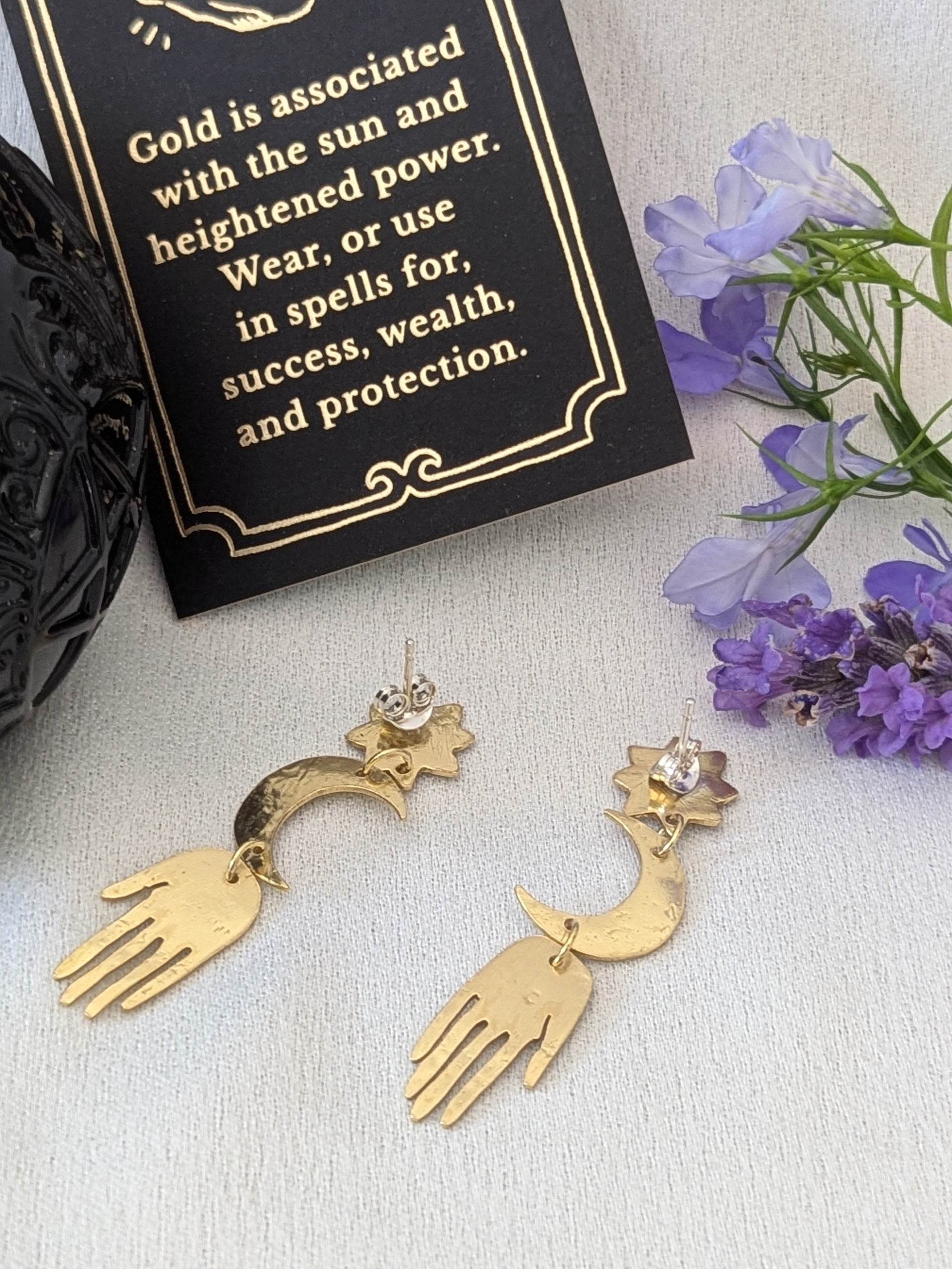 Gold brass earrings placed on a white background showing the back of the earrings. Hand-cut from raw gold brass, featuring a star stud, a crescent moon, and a protective hand delicately dangling below. In the background there is purple flowers next to a card that details the symbolism of wearing gold which is success, wealth and protection.
