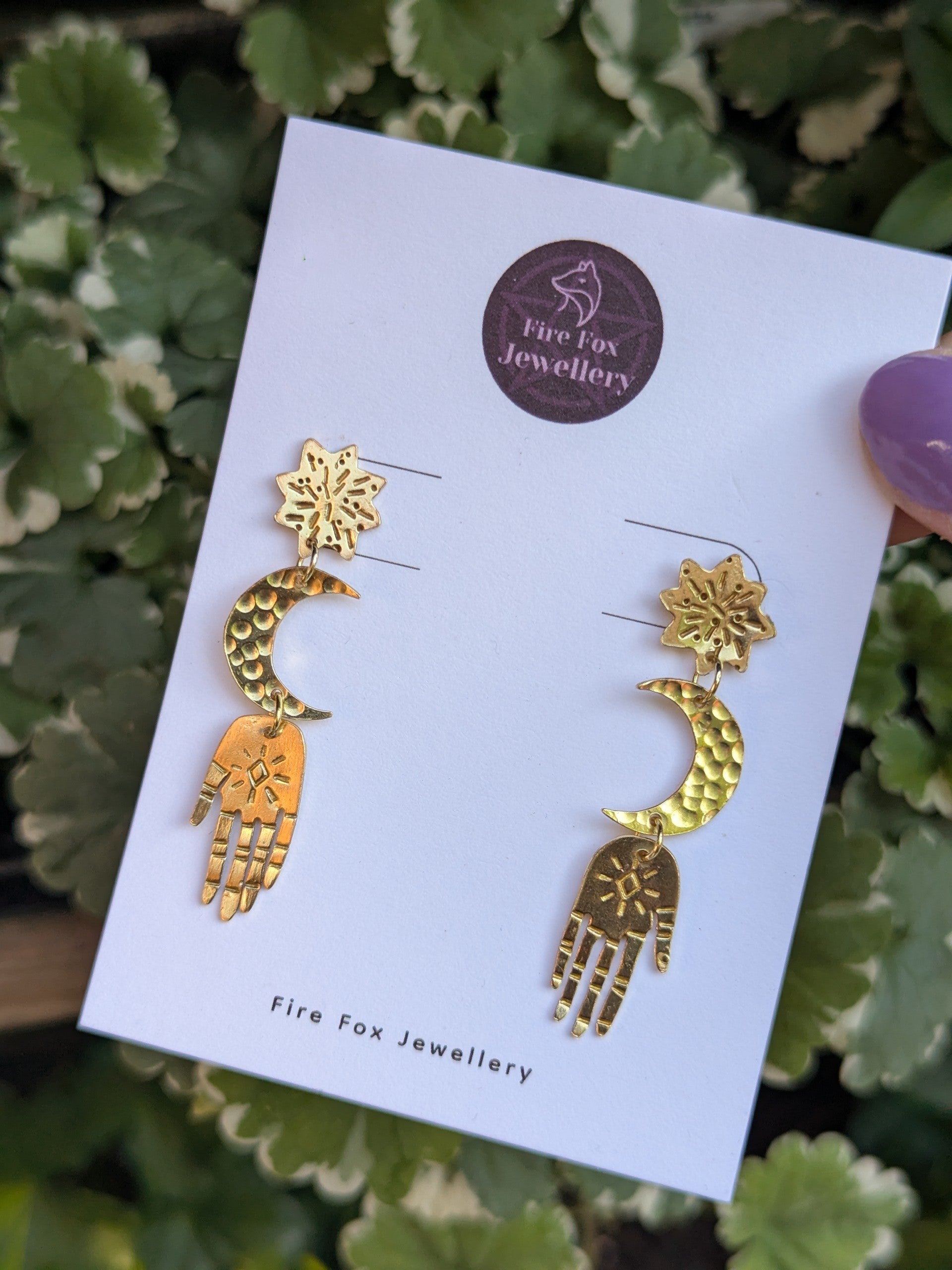 Gold brass earrings placed on a white Fire Fox Jewellery earring card to enhance the beautiful detail of each component. Hand-cut from raw gold brass, featuring a star stud, a crescent moon, and a protective hand delicately dangling below. There is a greenery in the background.
