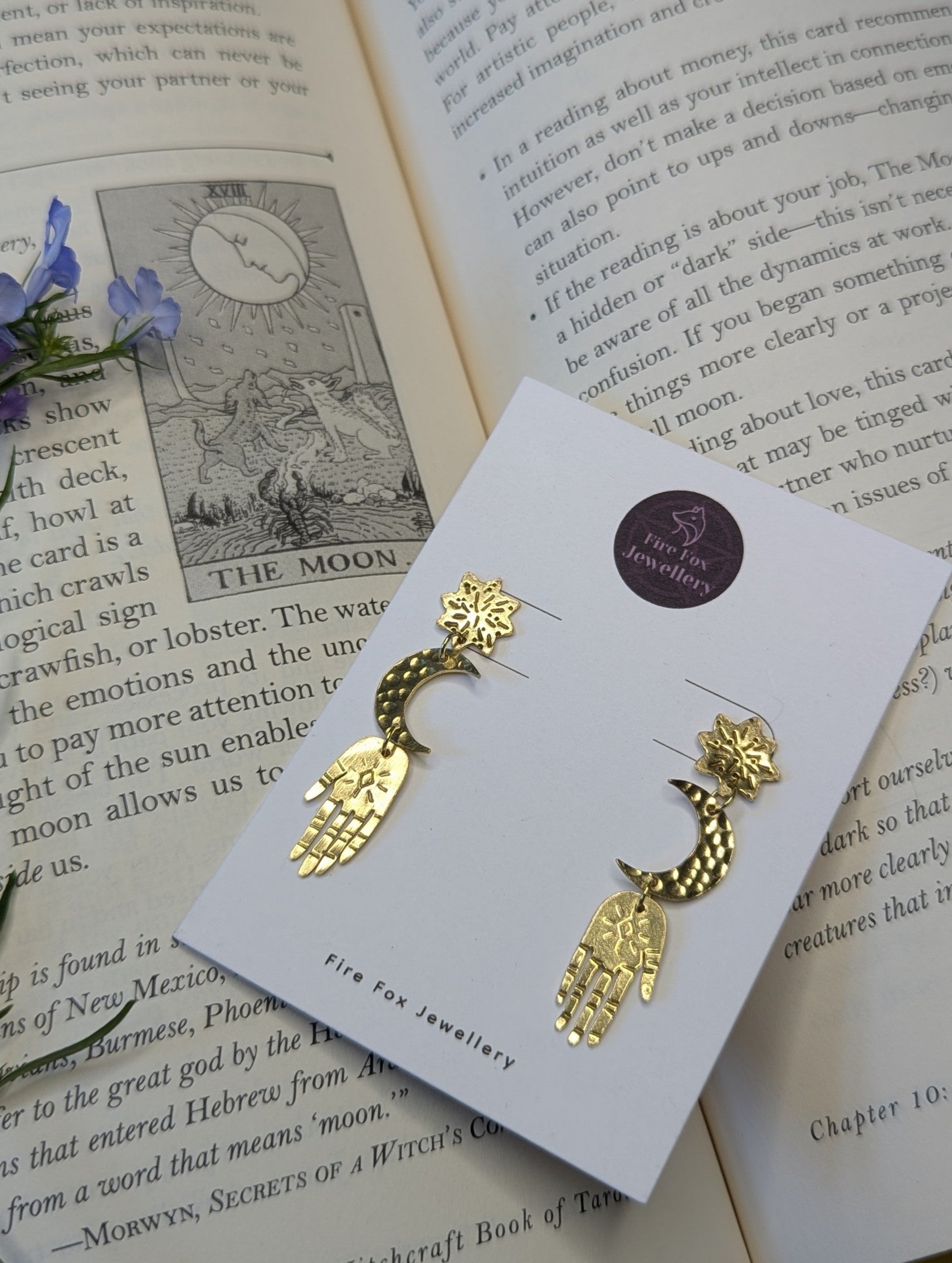 Gold brass earrings placed on a white Fire Fox Jewellery display card to enhance the beautiful detail of each component. Hand-cut from raw gold brass, featuring a star stud, a crescent moon, and a protective hand delicately dangling below. The earrings are placed on a open page of a tarot card book, on the page of the moon tarot card.