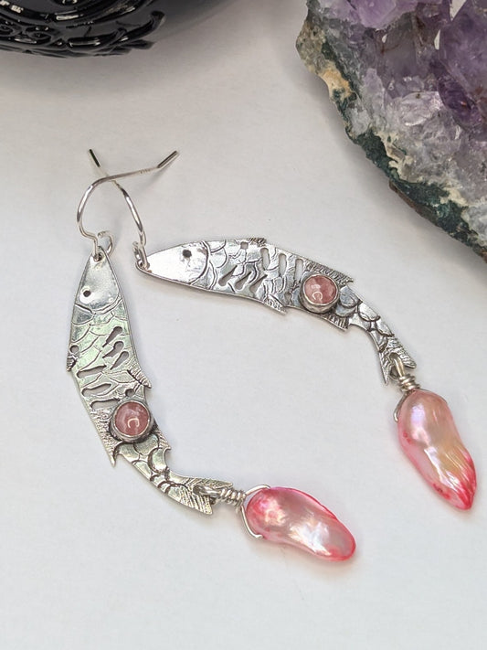 Sterling silver fish earrings, with fish details engraved on the silver, adorned with pink Rhodochrosite Cabochons and Pink keshi Freshwater Pearls. The earrings are placed on a white background with an amethyst stone and spell jar in the background.
