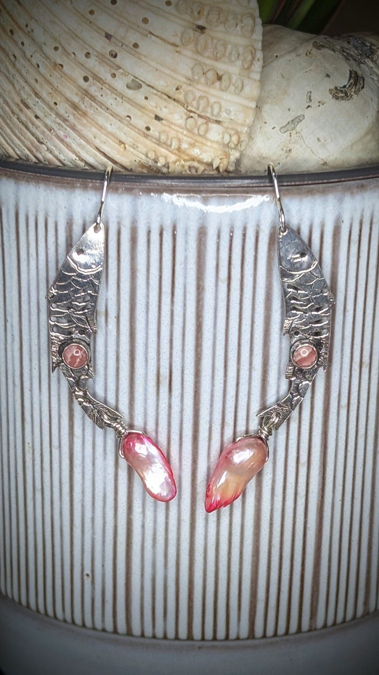 Sterling silver fish earrings, with fish details engraved on the silver, adorned with pink Rhodochrosite Cabochons and Pink keshi Freshwater Pearls. The earrings are hanging from a stripey plant with shells above the earrings. 