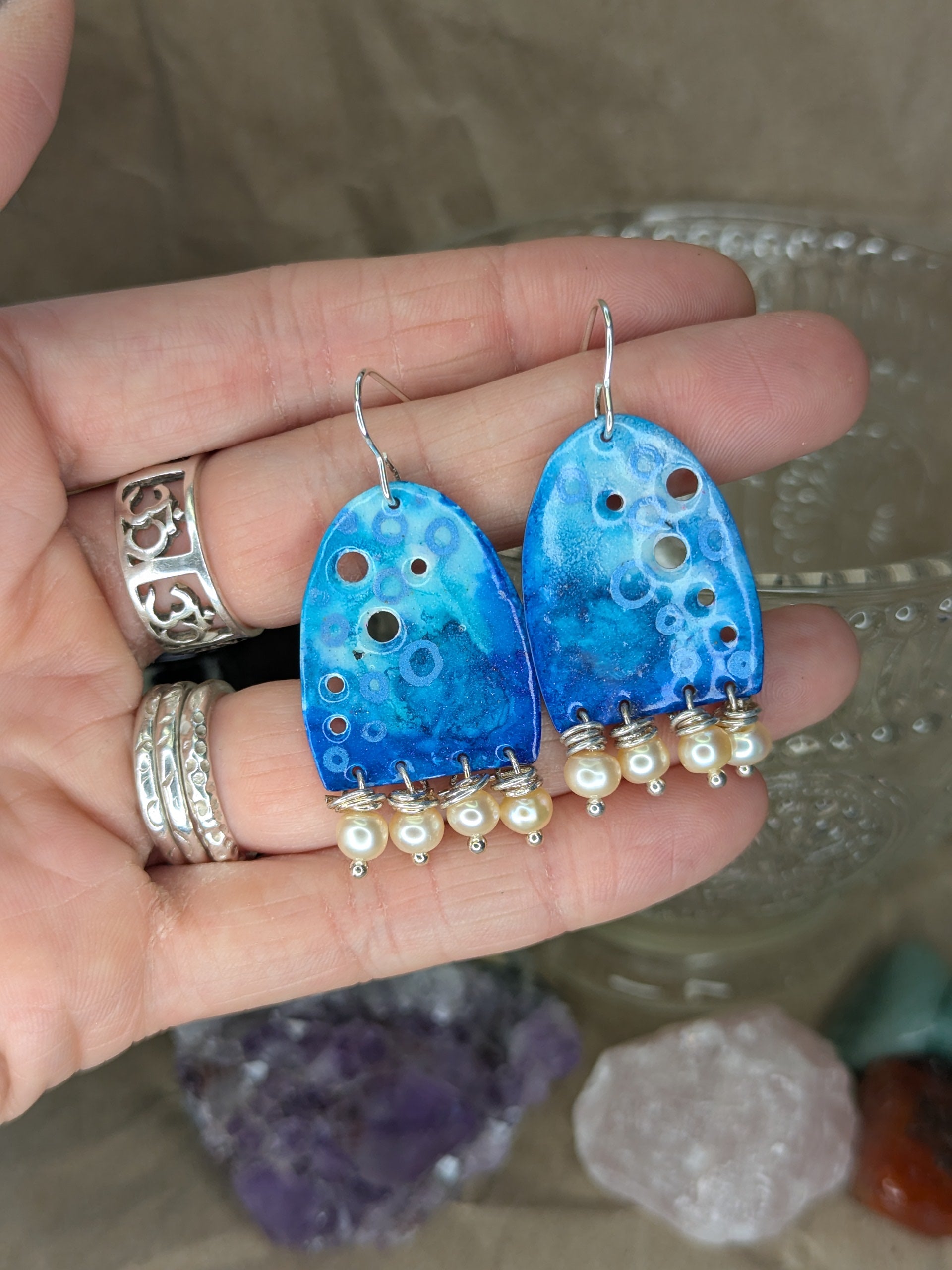 Blue enamel earrings with colours of blue with apricot freshwater pearls, Earrings are placed on hands to show the contrast with the earrings against skin.
