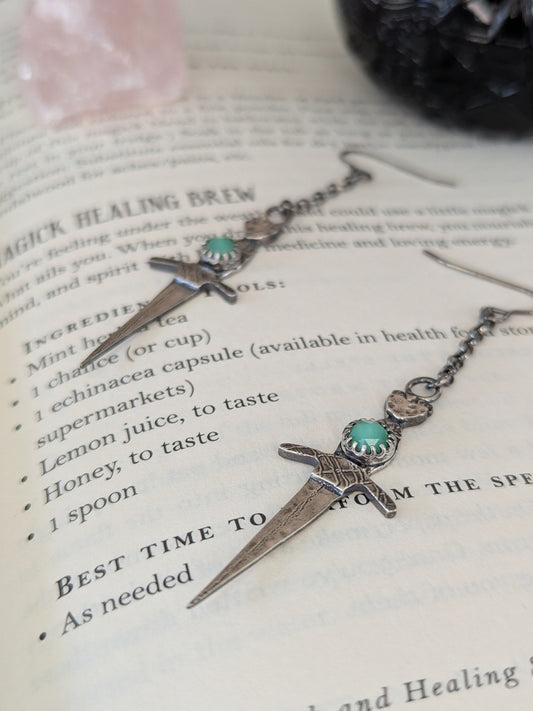 Athame witch's ritual dagger earrings displayed on the pages of an open spell book, showcasing their intricate design in recycled silver with chrysoprase stones and oxidised finish.