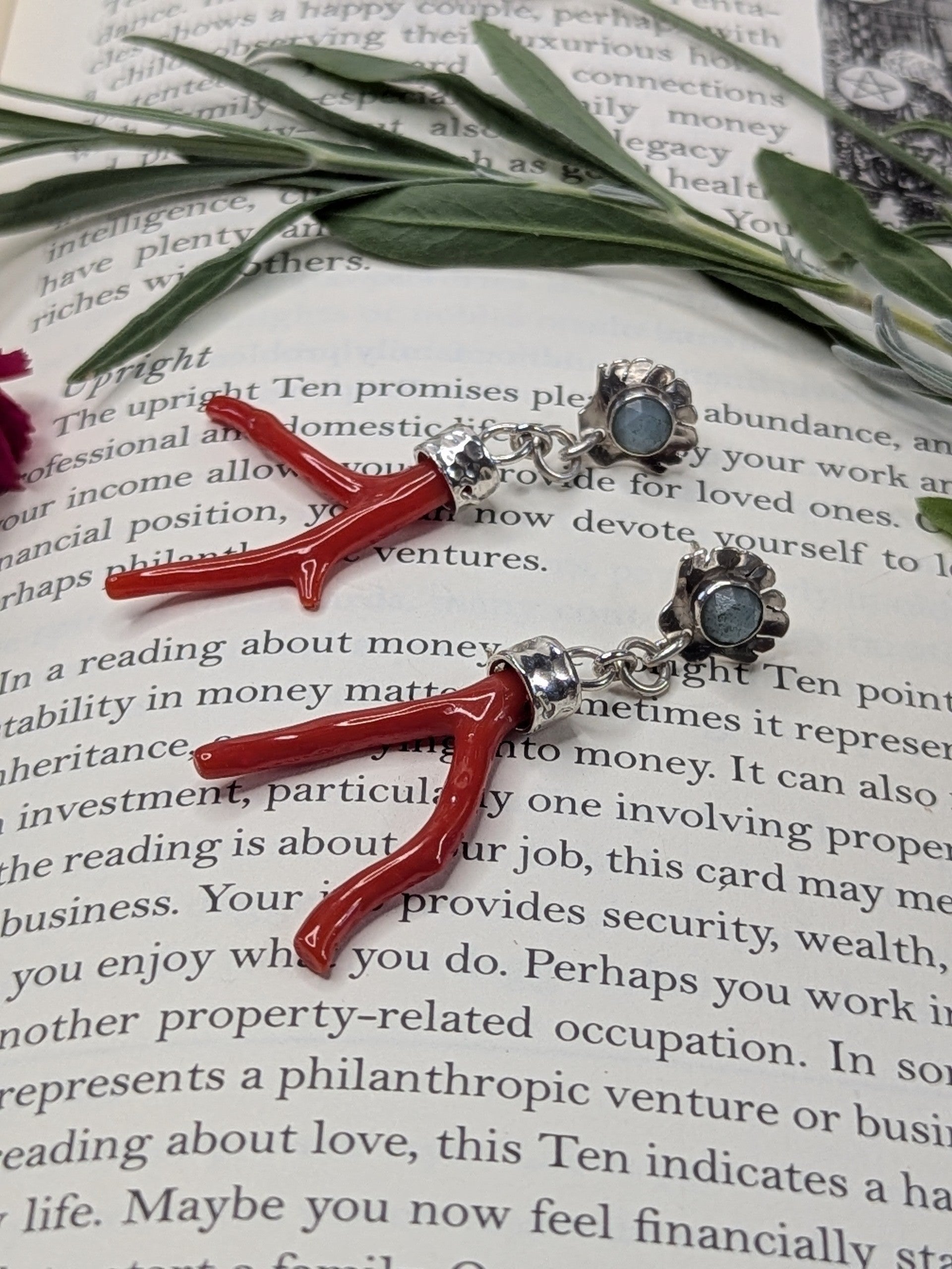 Sterling silver stud earrings styled on an open book, featuring a rose-cut turquoise stone set in scallop shell studs, with red coral branch drops hanging below. Surrounded by sprigs of lavender, sage, and a pink carnation flower, evoking a mystical and botanical aesthetic.