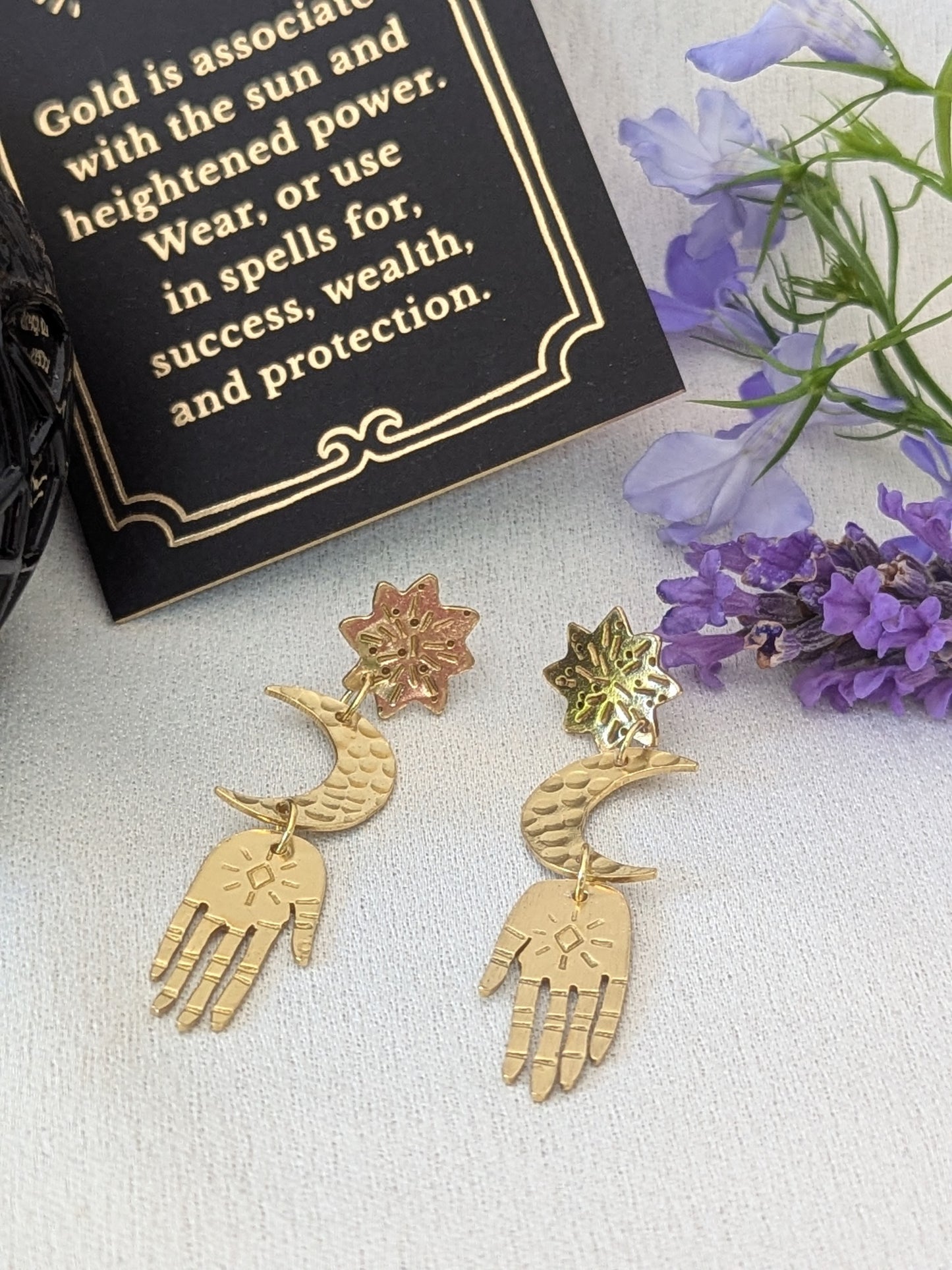 Gold brass earrings placed on a white background to enhance the beautiful detail of each component. Hand-cut from raw gold brass, featuring a star stud, a crescent moon, and a protective hand delicately dangling below. In the background there is purple flowers next to a card that details the symbolism of wearing gold which is success, wealth and protection.