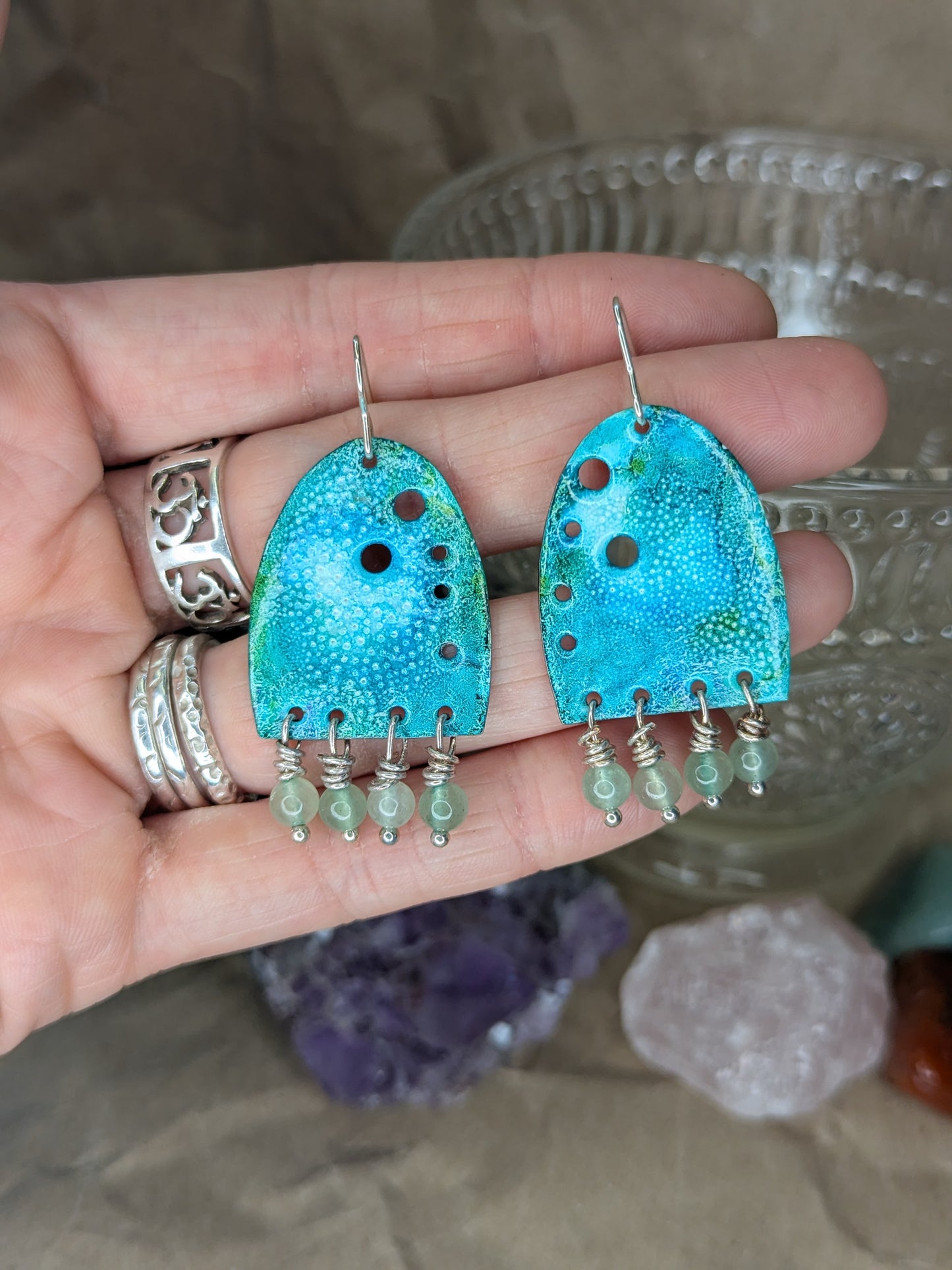 Turquoise enamel earrings with colours of blue and turquoise adorned with green aventurine beads, Earrings are placed on hands to show the contrast with the earrings against skin.

