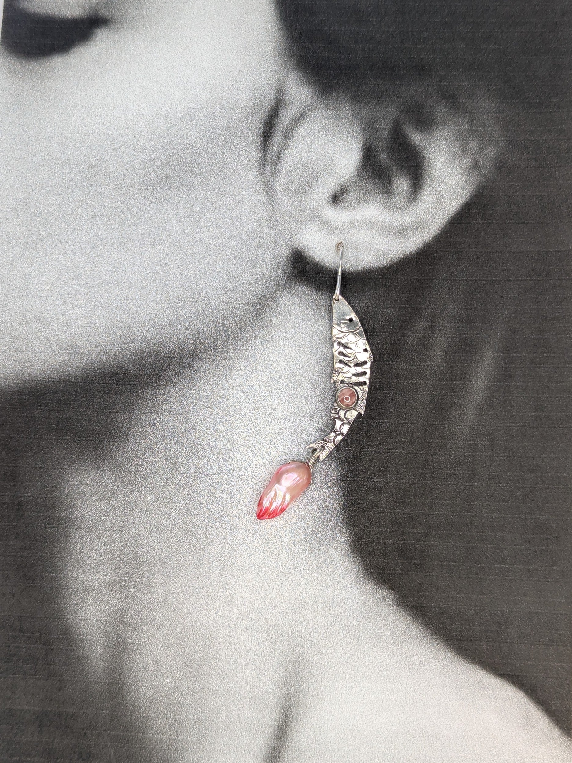 Sterling silver fish earrings, with fish details engraved on the silver, adorned with pink Rhodochrosite Cabochons and Pink keshi Freshwater Pearls. The earrings are placed on a black and white image of a model to show the earrings in the ear lobe. 
