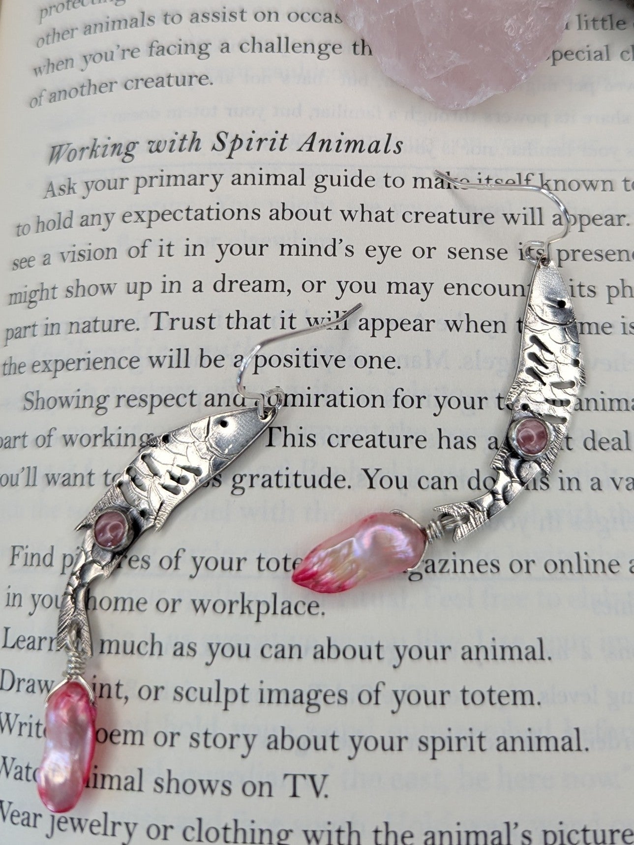 Sterling silver fish earrings, with fish details engraved on the silver, adorned with pink Rhodochrosite Cabochons and Pink keshi Freshwater Pearls. The earrings are placed on a spell book page that is detailing spiritual animals in magic. There is a rose quartz in the background. 