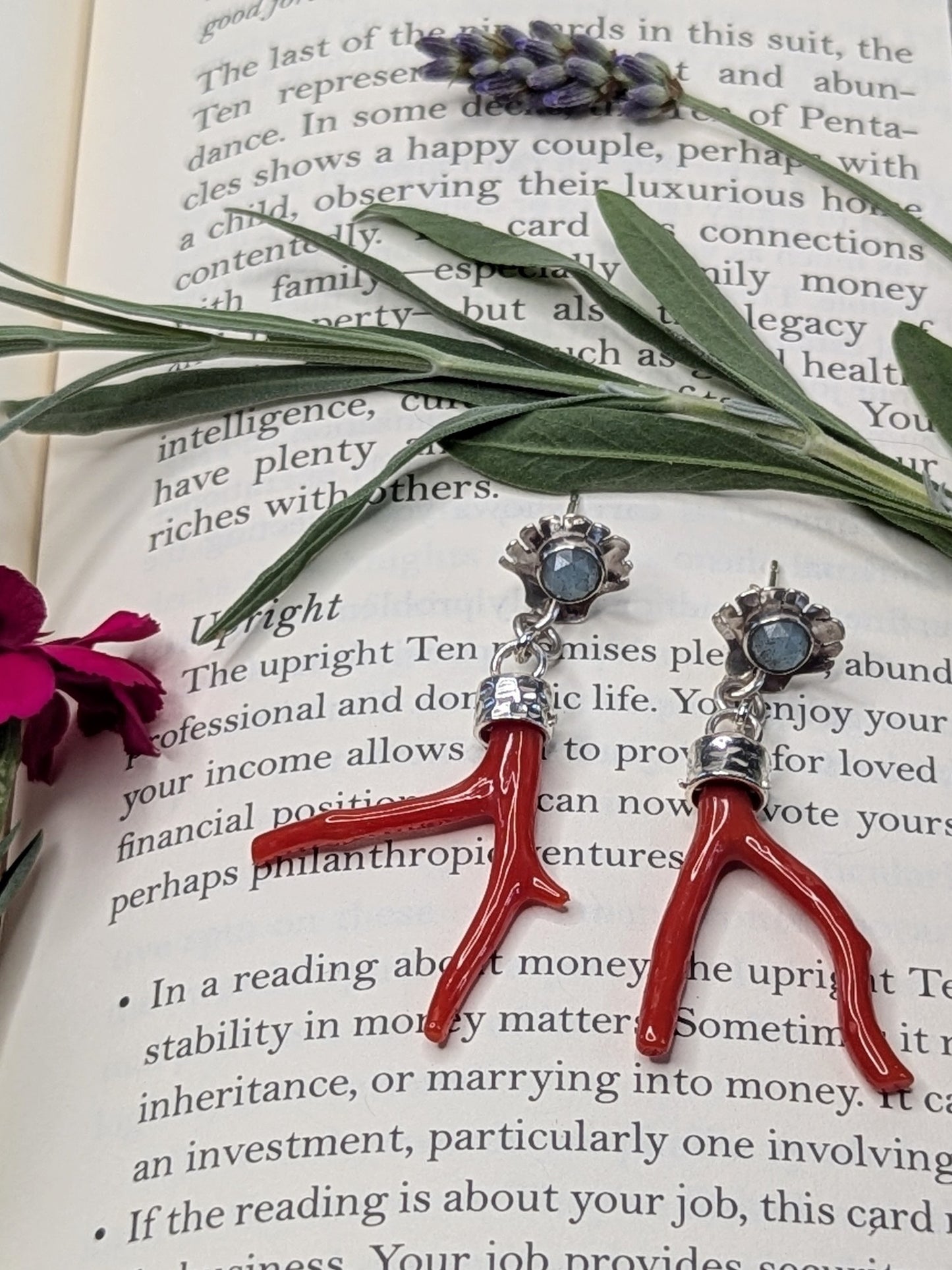 Sterling silver stud earrings styled on an open book, featuring a rose-cut turquoise stone set in scallop shell studs, with red coral branch drops hanging below. Surrounded by sprigs of lavender, sage, and a pink carnation flower, evoking a mystical and botanical aesthetic.