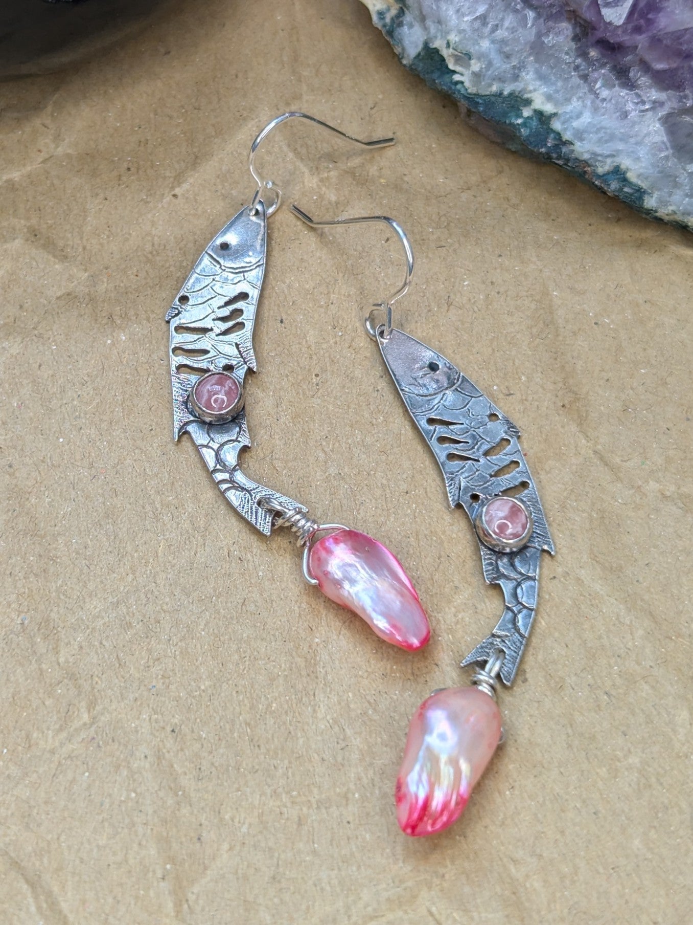 Sterling silver fish earrings, with fish details engraved on the silver, adorned with pink Rhodochrosite Cabochons and Pink keshi Freshwater Pearls. The earrings are placed on a brown background with an amethyst stone in the background. 