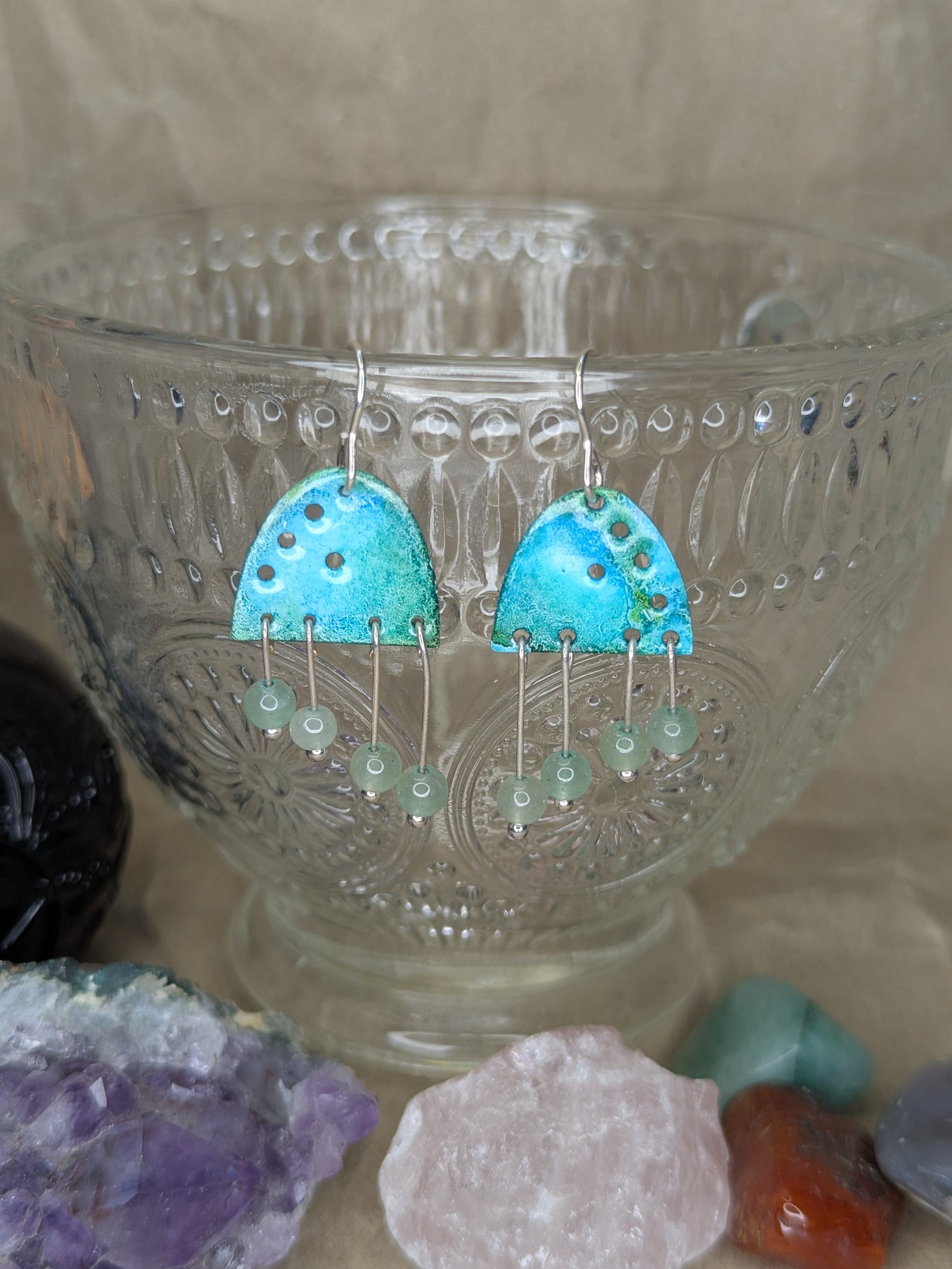 Handcrafted turquoise enamel earrings with colours of blue and turquoise adorned with green aventurine beads. The earrings are hanging from a crystal teacup
